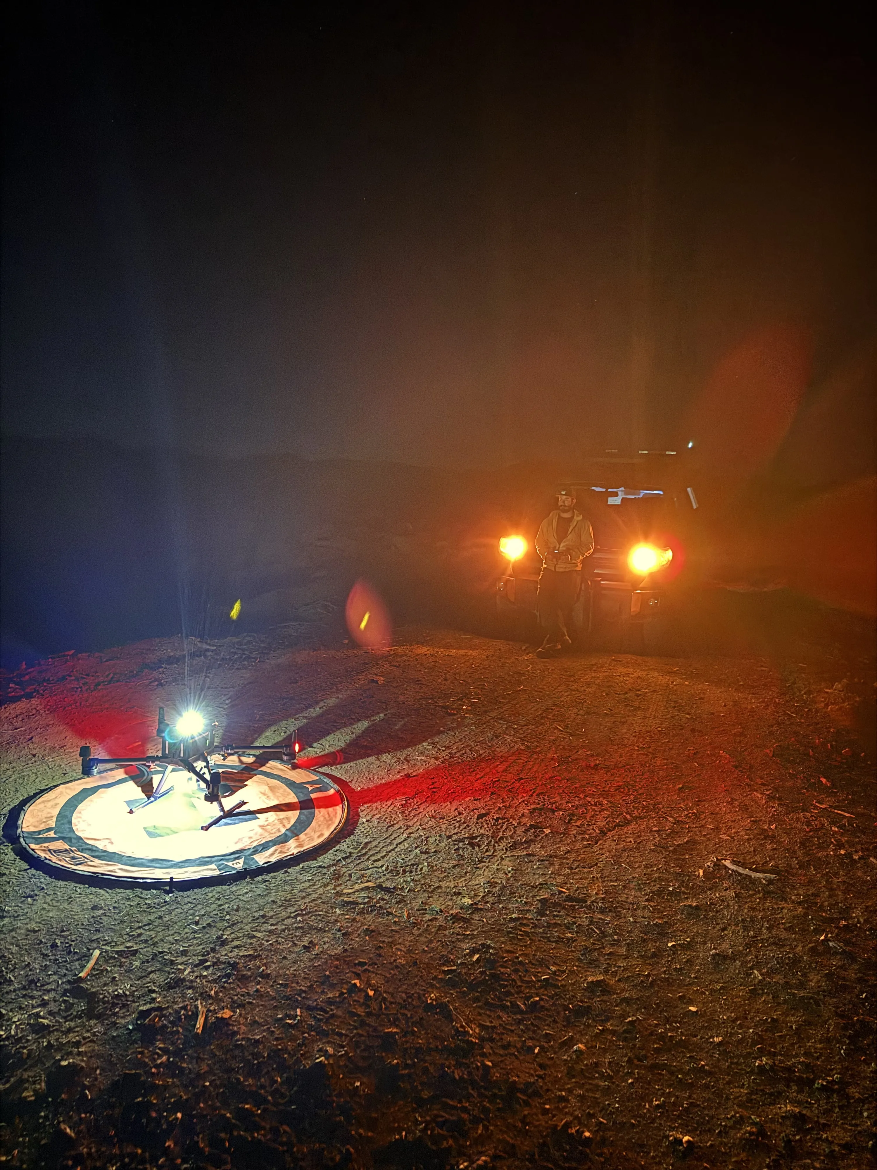 The aircraft on its landing pad at night, lit by headlamp, with the truck lights glowing behind.