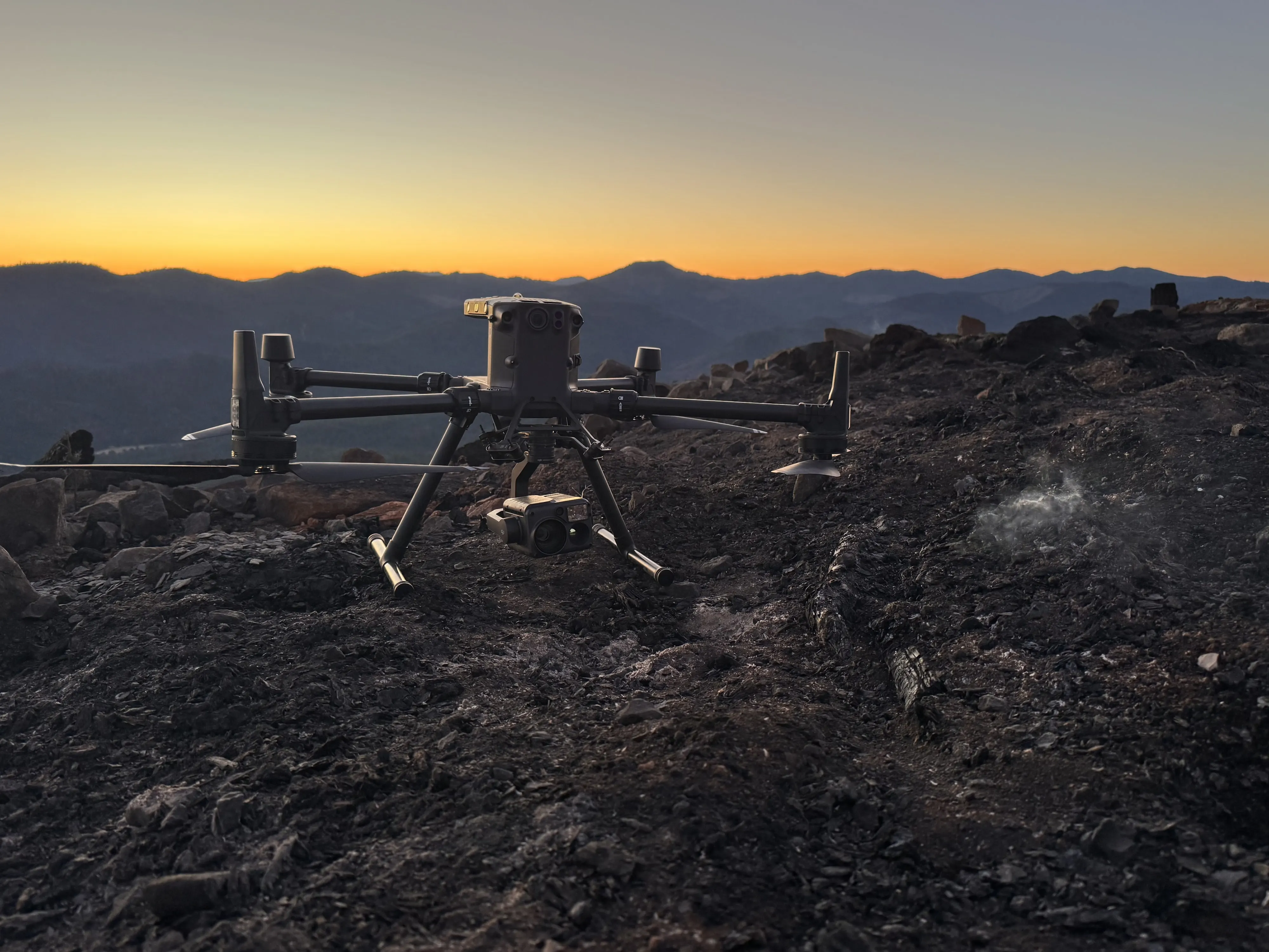 Matrice 300 with a thermal camera sitting on a burned ridge at sunset, smoke drifting from the ash behind it.