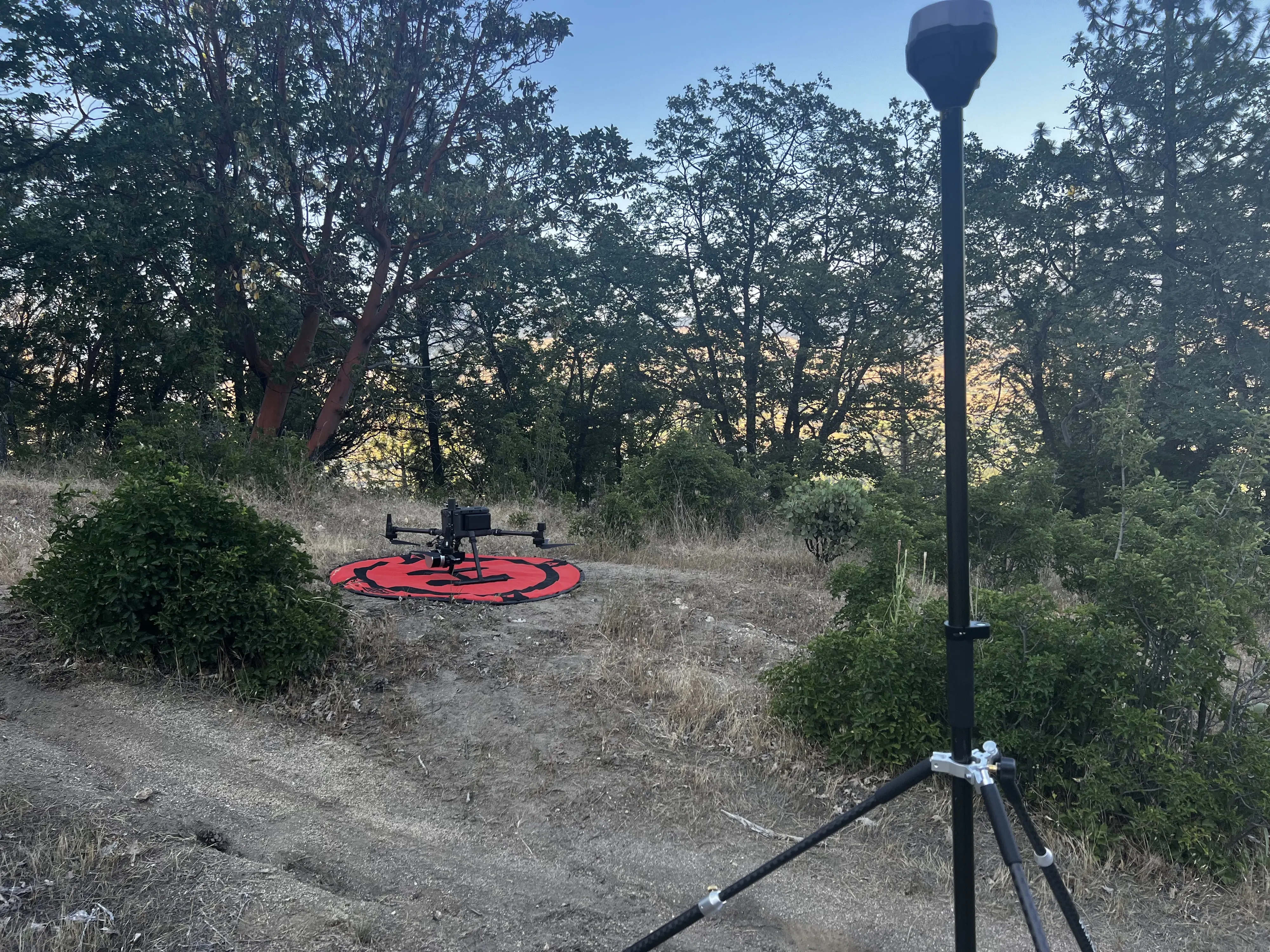 A GNSS base station on a tripod in the foreground and the Matrice 300 on its launch pad behind it, in a dry field under oaks.