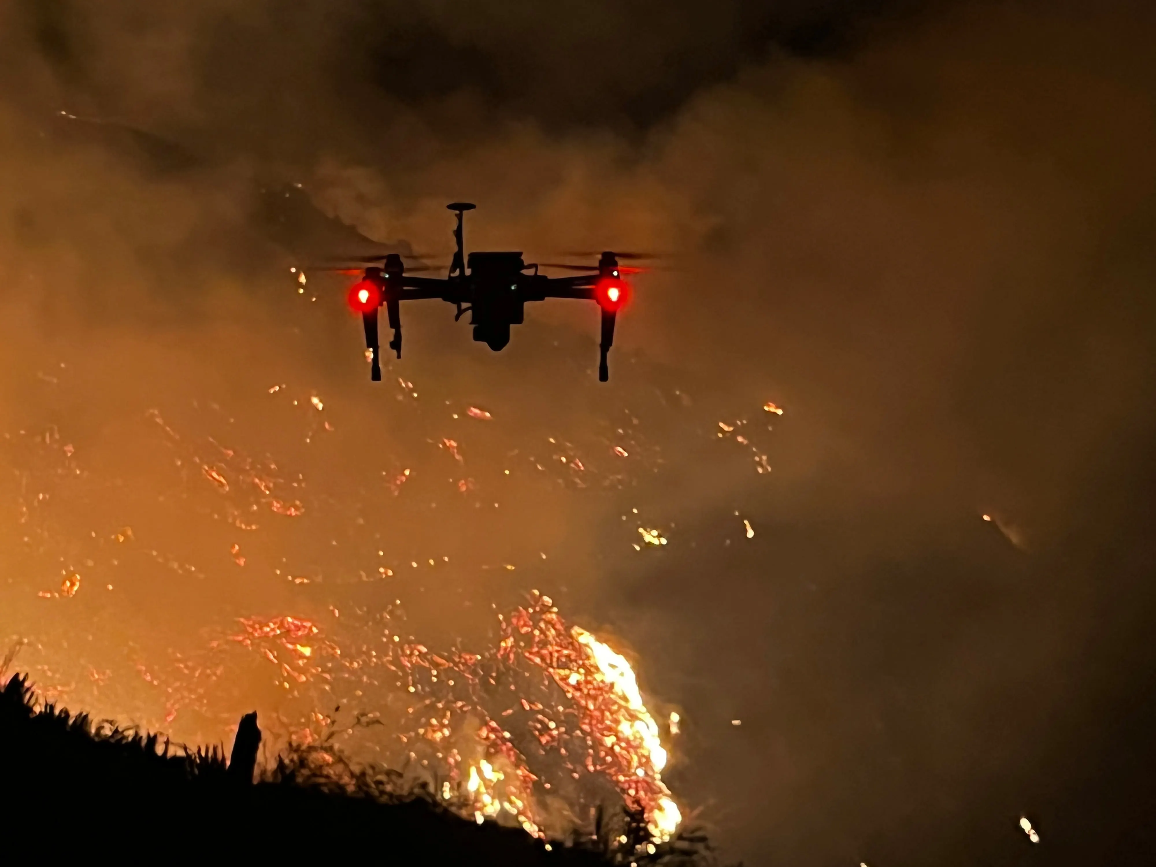 The aircraft hovering at night in front of a hillside of open flame and drifting embers.