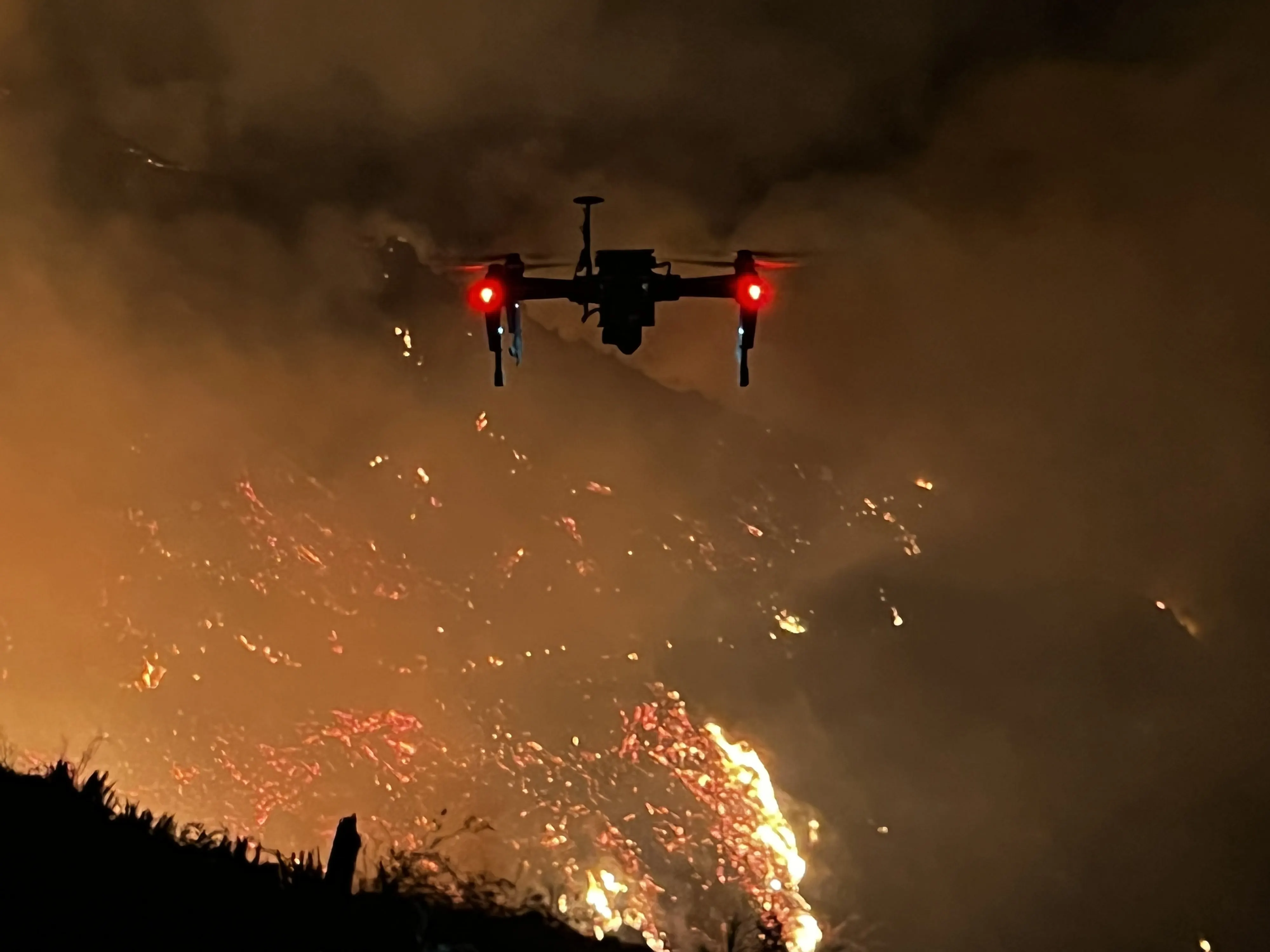 The aircraft hovering at night with its red lights on, a hillside of open flame and glowing embers behind it.